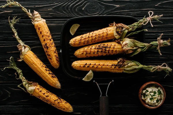 Vue du dessus de la plaque de cuisson avec du maïs grillé avec des tranches de citron vert et de la crème avec du persil sur une table en bois — Photo de stock