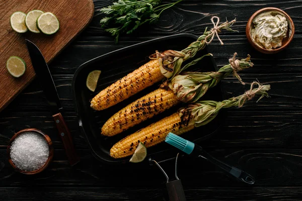 Vue du dessus de la plaque de cuisson avec du maïs grillé et des tranches de lime entourées de sel et de crème avec du persil sur une table en bois — Photo de stock