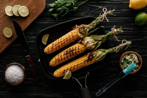 Vue du dessus de la plaque de cuisson avec du maïs grillé et des tranches de lime entourées d'ingrédients sur une table en bois — Photo de stock