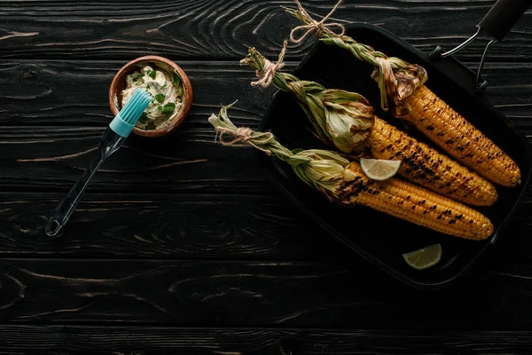 Vue du dessus de la plaque de cuisson avec du maïs grillé avec des tranches de citron vert, brosse de cuisson et beurre avec du persil sur une table en bois — Photo de stock