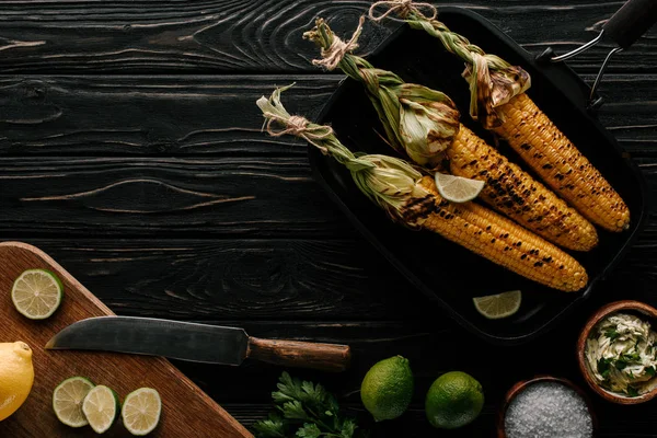 Top view of grilled corn, cutting board with lemon and lime slices with knife on wooden table — Stock Photo