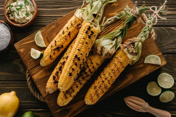 Top view of cutting board with grilled corn, lime slices, salt, lemon, butter with parsley and squeezer on wooden table — Stock Photo