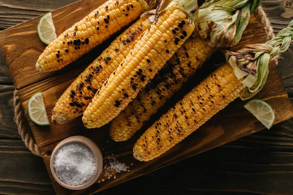 Top view of cutting board with delicious grilled corn, lime slices and salt on wooden table — Stock Photo