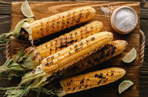 Draufsicht auf Schneidebrett mit gegrilltem Mais, Limettenscheiben und Salz auf Holztisch — Stockfoto