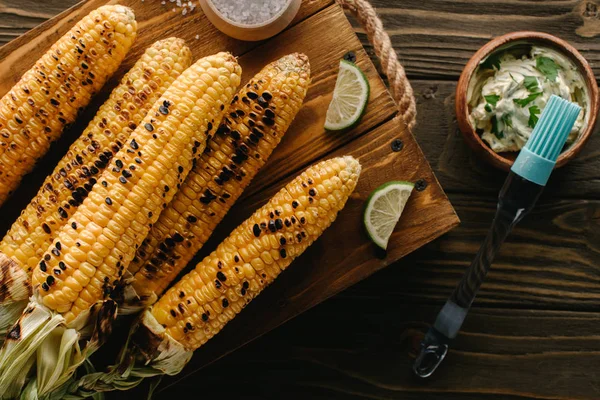 Top view of cutting board with grilled corn, lime slices near cooking brush and butter with parsley on wooden table — Stock Photo