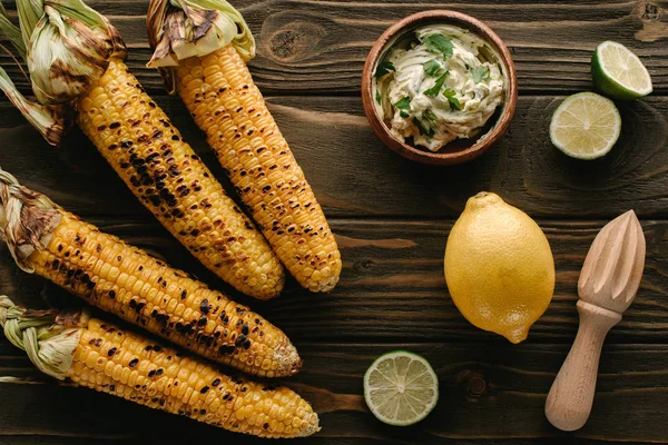 Top view of grilled corn, lime slices, lemon, wooden squeezer and cream with parsley on wooden table — Stock Photo