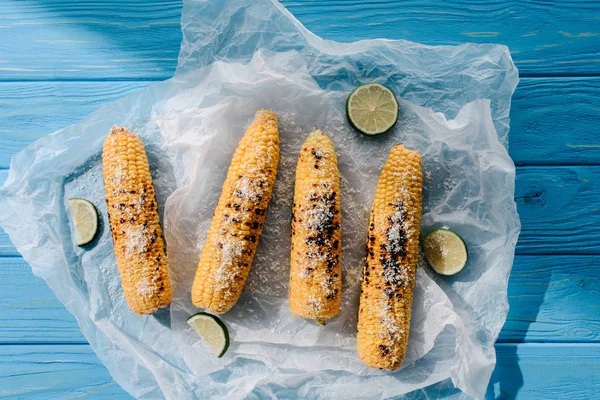 Vue surélevée du maïs salé grillé avec des tranches de lime sur du papier cuisson sur une table en bois — Photo de stock