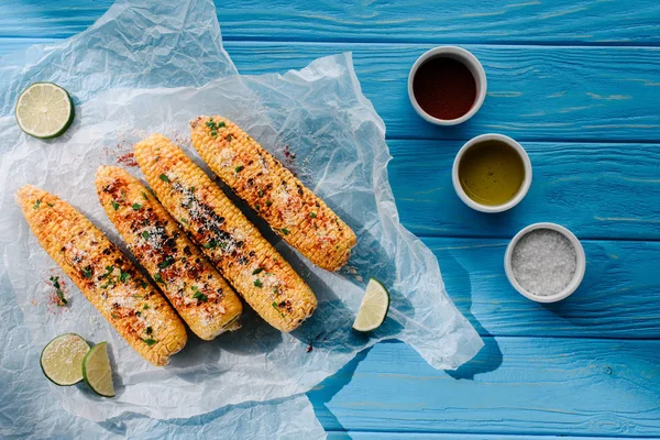 Vue du dessus du papier cuisson avec du maïs grillé près d'une épice chili, de l'huile et du sel sur une table en bois — Photo de stock