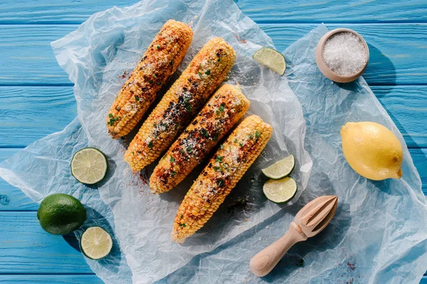 Top view of grilled corn on baking paper with lime slices, lemon, salt and wooden squeezer on table — Stock Photo