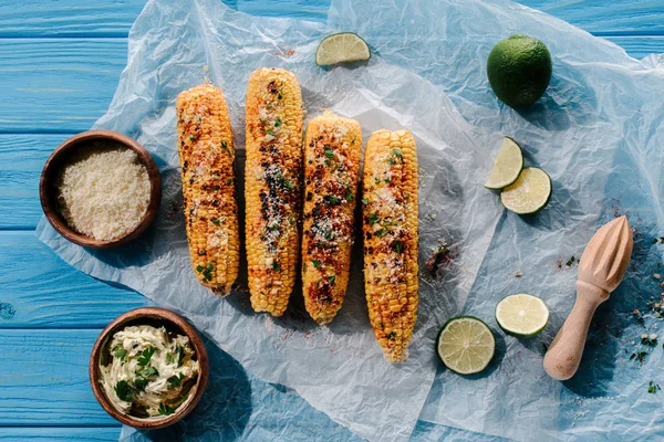 Top view of grilled corn on baking paper with lime slices, squeezer, cream with parsley and spice — Stock Photo