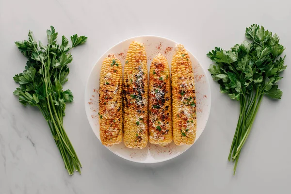 Top view of grilled corn with salt and chili spice on plate near parsley on marble table — Stock Photo