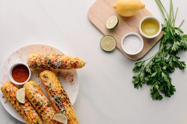 Ansicht von gegrilltem Mais, Limettenscheiben und Chili-Gewürz auf Teller neben Schneidebrett mit Salz, Öl, Zitrone und Petersilie auf Marmortisch — Stockfoto