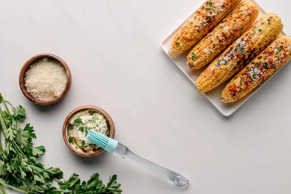 Vue de dessus de la plaque avec du maïs grillé, épices, brosse de cuisson et beurre avec du persil sur table en marbre — Photo de stock