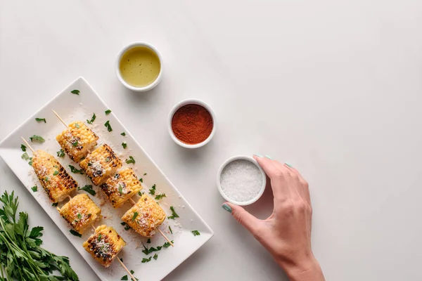 Cropped image of woman taking salt from marble table with grilled corn on plate near parsley — Stock Photo