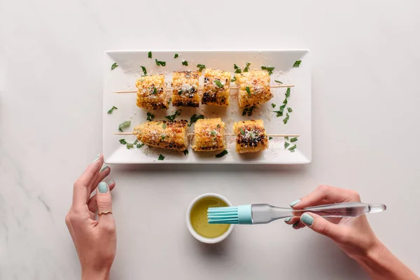 Cropped image of woman putting cooking brush in oil for spreading grilled corn on marble table — Stock Photo