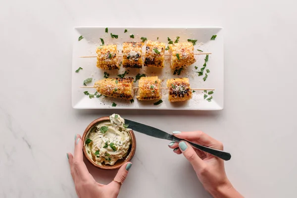 Cropped image of woman taking cream with parsley by knife over marble table with grilled corn on plate — Stock Photo