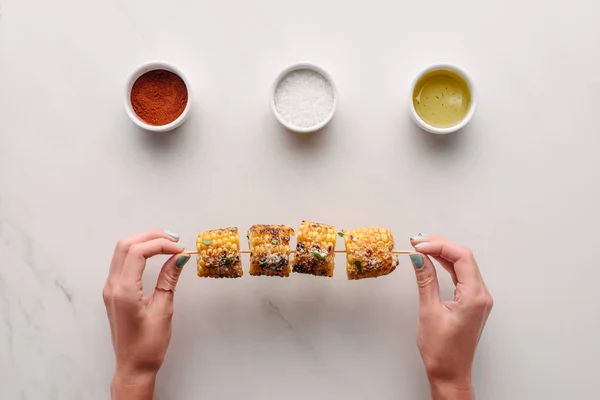 Imagen recortada de la mujer sosteniendo maíz a la parrilla sobre la mesa de mármol con sal, aceite y especias de chile - foto de stock