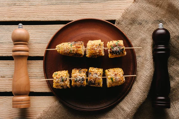 Top view of plate with grilled corn between salt and pepper grinders on wooden table with sackcloth — Stock Photo