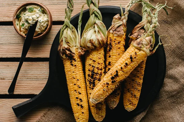 Top view of grilled corn, knife and butter with parsley on wooden table with sackcloth — Stock Photo