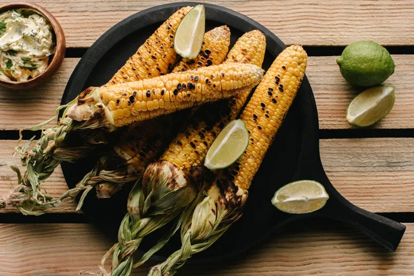 Вид вкусной кукурузы на гриле с ломтиками лайма рядом с маслом с петрушкой на деревянном столе — стоковое фото