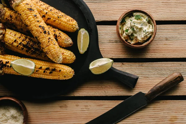 Vista dall'alto di mais alla griglia, fette di lime, coltello e burro con prezzemolo sul tavolo di legno — Foto stock