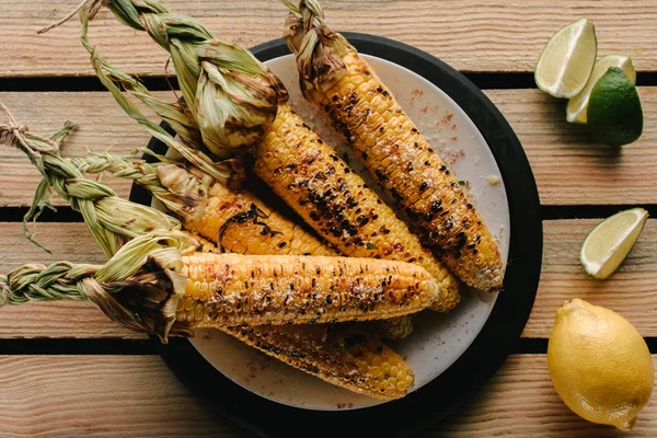 Vue de dessus de délicieux maïs grillé sur assiette et tranches de citron vert sur table en bois — Photo de stock