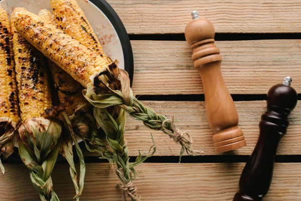 Вид жареной кукурузы на тарелке рядом с солью и перцовыми шлифовальными станками на деревянном столе — стоковое фото