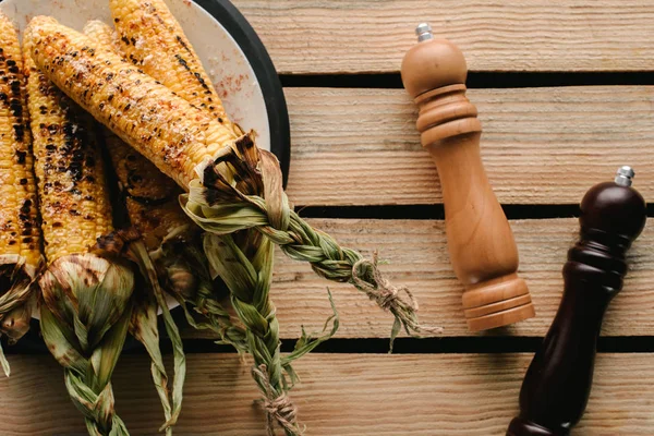 Top view of grilled corn on plate near salt and pepper grinders on wooden table — Stock Photo