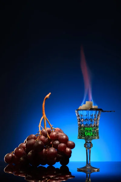 Vaso de absenta en llamas con azúcar y uvas sobre fondo azul oscuro — Stock Photo