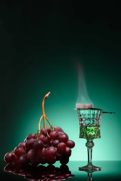 Vaso de absenta ardiente con azúcar y uvas sobre fondo azul oscuro - foto de stock