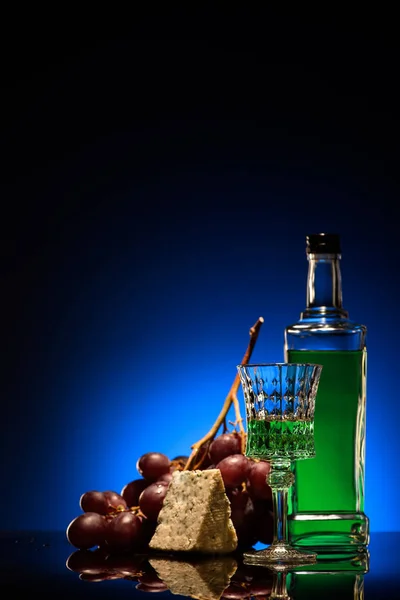 Nahaufnahme von Absinth mit Trauben und Dorblu-Käse auf Spiegeloberfläche auf dunkelblauem Hintergrund — Stockfoto