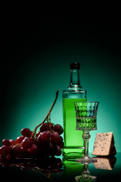 Nahaufnahme von Absinth mit Trauben und Käse auf Spiegeloberfläche auf dunkelblauem Hintergrund — Stockfoto