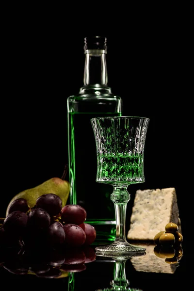 Close-up shot of absinthe with ripe fruits and cheese on mirror surface on black — Stock Photo