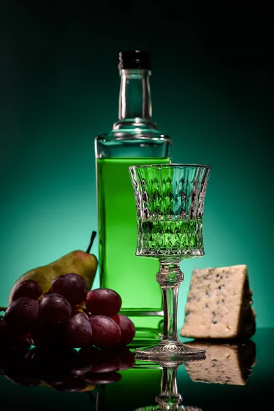 Close-up shot of absinthe with ripe fruits and cheese on mirror surface on dark blue background — Stock Photo