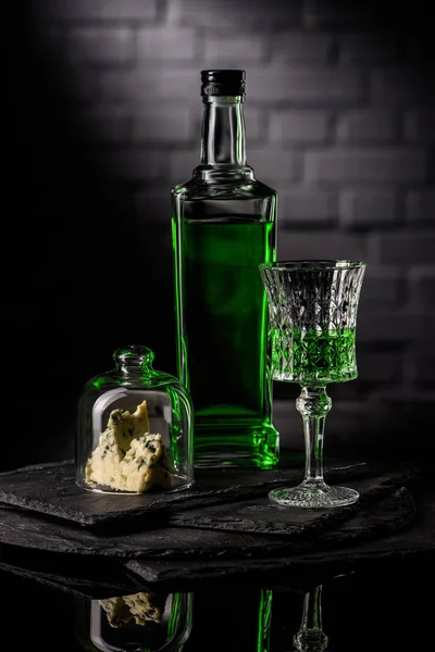 Close-up shot of glass and bottle of absinthe with slice of cheese on dark brick wall background — Stock Photo