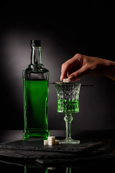 Tiro cortado de mulher colocando cubo de açúcar na colher absinto em vidro no fundo escuro — Fotografia de Stock