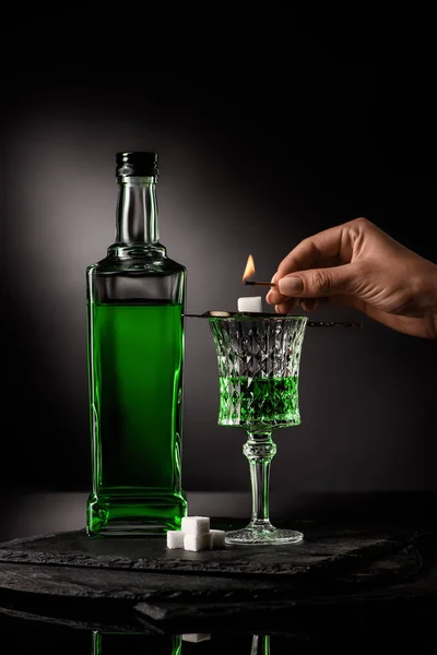 Abgeschnittene Aufnahme einer Frau, die Streichholz über Löffel mit Zuckerwürfel auf Absinthglas auf dunklem Hintergrund hält — Stockfoto