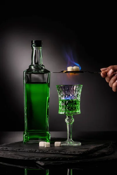 Cropped shot of woman holding spoon with sugar cubes over burning absinthe in glass on dark background — Stock Photo