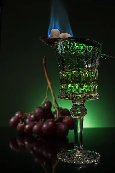 Lead glass of absinthe with branch of grapes and burning sugar on spoon on reflective surface and dark green background — Stock Photo