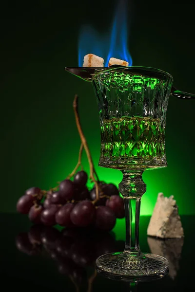 Crystal glass of absinthe with branch of grapes and burning sugar on spoon on reflective surface and dark green background — Stock Photo