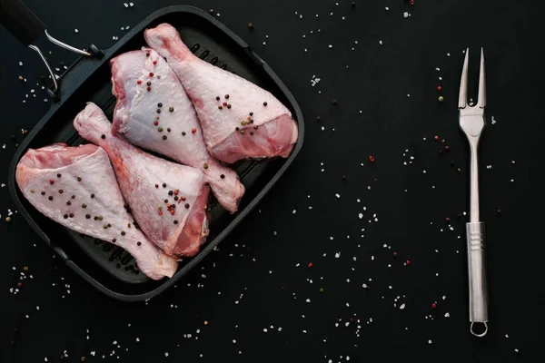 Ansicht von rohen Hühnerkeulen mit Pfefferkörnern und Salz in Grillpfanne mit Fleischgabel auf schwarz — Stockfoto