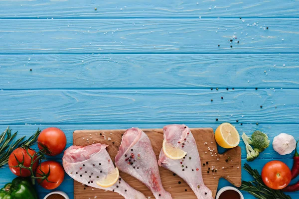 Vista superior de pernas de frango na tábua de madeira com diferentes vegetais na mesa azul — Fotografia de Stock