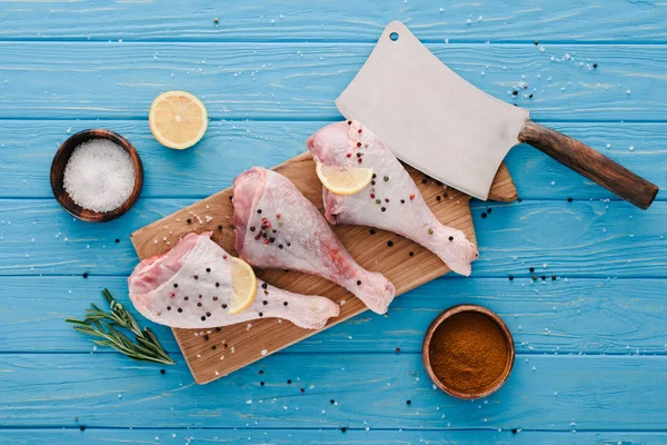 Vue du dessus des cuisses de poulet crues sur une planche de bois avec des épices et un coupe-plat bleu — Photo de stock