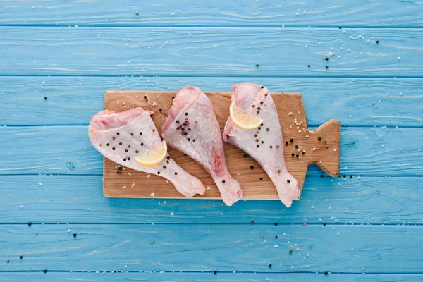 Vista superior de patas de pollo crudo con granos de pimienta y limón sobre tabla de madera sobre mesa azul - foto de stock