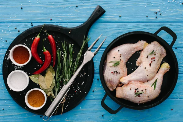 Vista superior de las patas de pollo en la sartén y diferentes especias en la tabla de cortar con tenedor de carne en la mesa azul - foto de stock