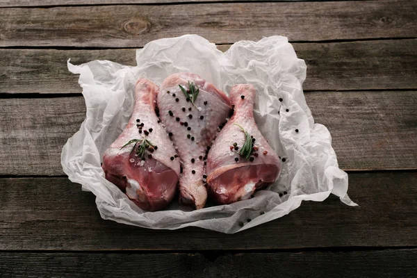 Raw turkey legs with pepper corns and rosemary on baking paper on wooden tabletop — Stock Photo