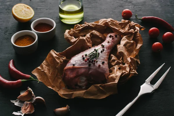 Una pata de pavo con granos de pimienta sobre papel de hornear sobre una mesa de madera con verduras y especias - foto de stock