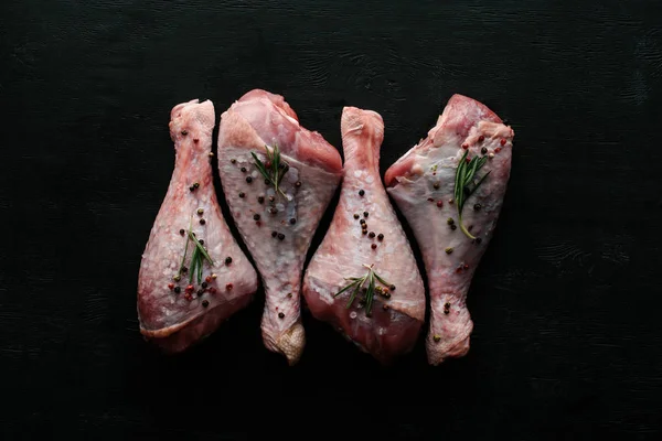 Vue du dessus des cuisses de poulet crues avec des grains de poivre et du romarin sur plateau en bois noir — Photo de stock