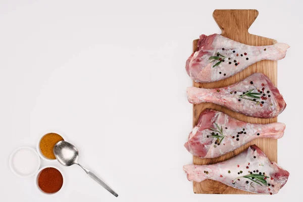 Vue du dessus des cuisses de poulet avec romarin sur planche à découper et épices dans des bols en bois, isolé sur blanc — Photo de stock