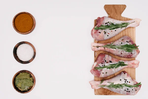 Vue du dessus des cuisses de poulet crues avec romarin sur planche de bois avec sel, paprika et feuilles de laurier dans des bols, isolé sur blanc — Photo de stock